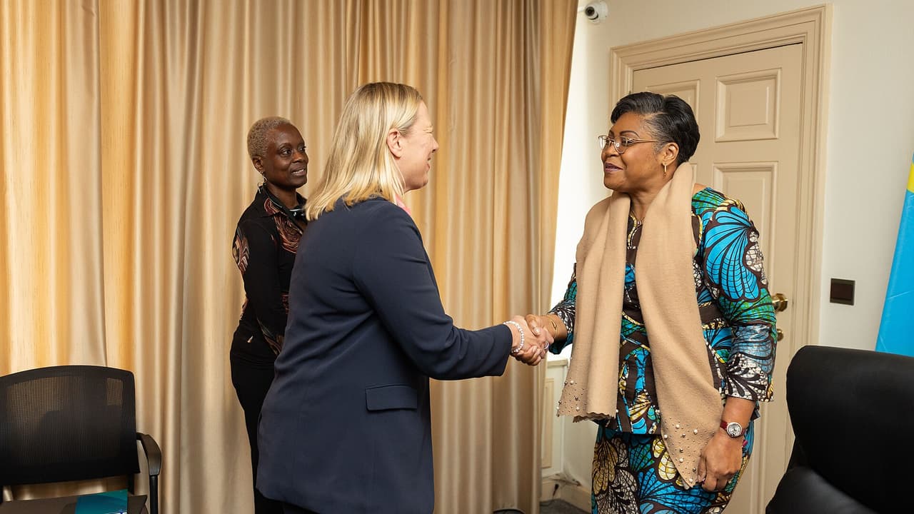 La Première ministre congolaise Judith Suminwa, à droite, serre la main de la commissaire européenne Jutta Urpilainen dans un salon de l'ambassade de la RDC à Bruxelles. Le drapeau congolais est visible à l'arrière-plan.
