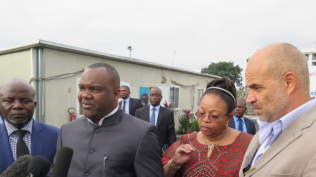 Corneille Nangaa, vêtu d'une veste grise à col mao, répond aux questions de journalistes qui lui tendent des micros. Il est entouré de plusieurs personnes, dont une femme en pagne rouge et un homme en veste beige portant un badge des Nations unies. En arrière-plan, des bâtiments préfabriqués.