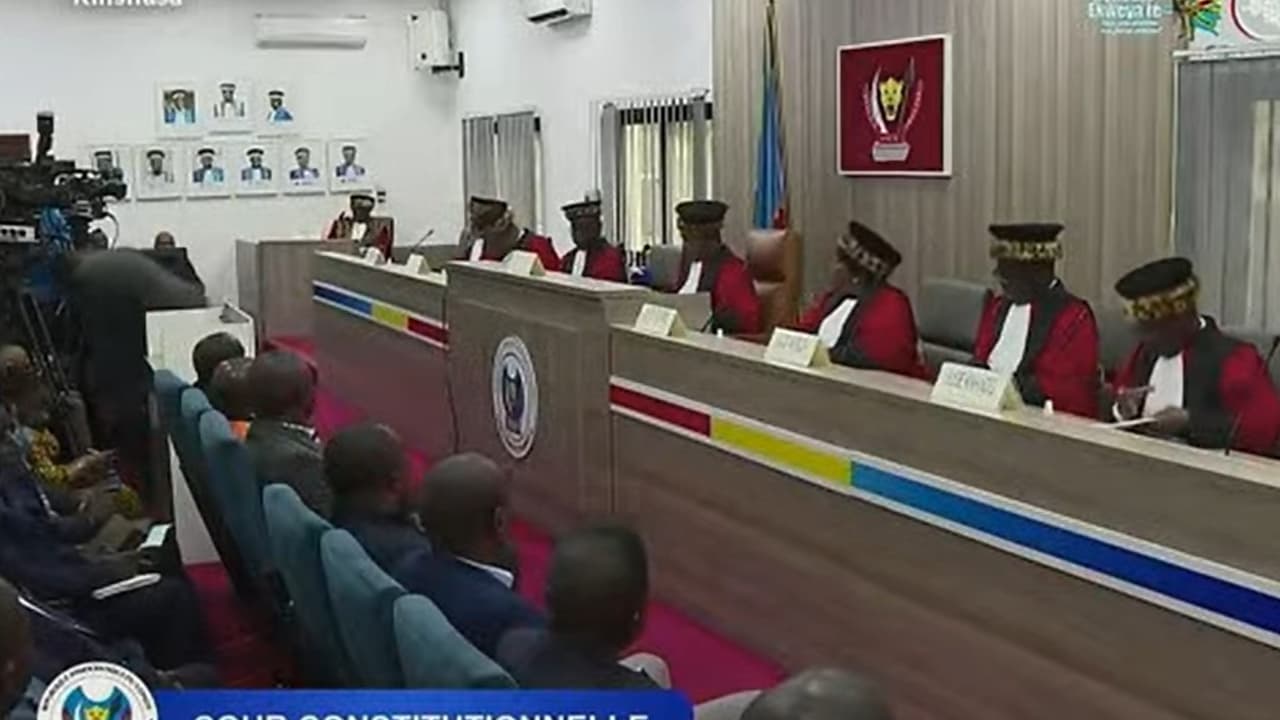 Les juges de la Cour constitutionnelle de la RDC, en toge rouge et toque bordée de peau de léopard, siègent en audience publique à Kinshasa, devant les armoiries de la République ; au premier plan, l'assistance et les caméras.