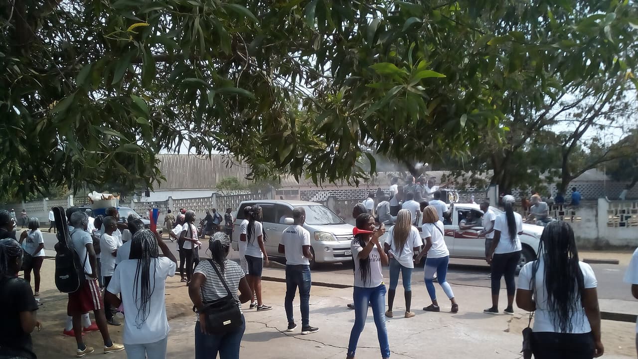 Des jeunes gens en t-shirts blancs rassemblés dans une rue de Kinshasa ; une jeune femme souffle dans une vuvuzela, d'autres sont montés à l'arrière d'une camionnette, sous des manguiers.