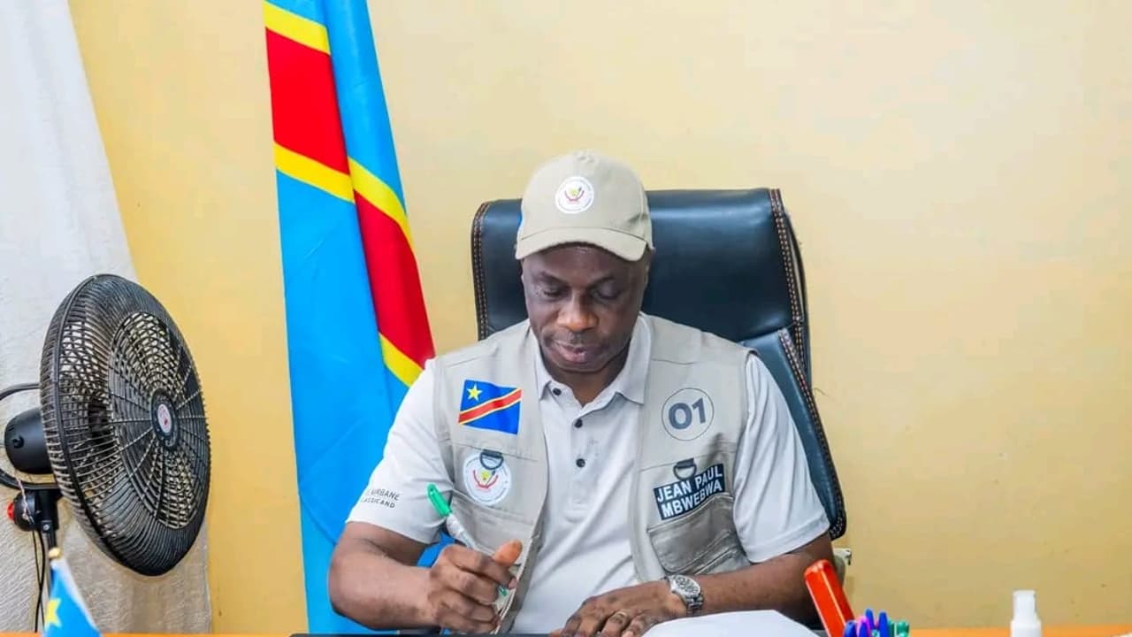 Le gouverneur du Kasaï-Oriental Jean-Paul Mbwebwa, casquette et gilet gris floqué de son nom et du numéro 01, signe un document à son bureau devant le drapeau de la RDC.