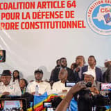 Les responsables de la Coalition Article 64 assis derrière une table drapée du drapeau congolais, devant une grande banderole blanche portant en lettres rouges « Coalition Article 64 pour la défense de l'ordre constitutionnel » et le logo C64 ; au premier plan, des caméras et des téléphones filment la séance.