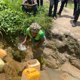 Une femme accroupie remplit des bidons jaunes avec une cuvette en puisant l'eau d'une source non aménagée creusée à même la terre, entourée de personnes debout au bord du trou.