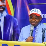 Ferdinand Kambere, secrétaire permanent adjoint du PPRD, parle au micro derrière un pupitre, coiffé d'une casquette blanche siglée P.P.R.D, devant une affiche à l'effigie de Joseph Kabila et une banderole du parti