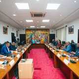 Vue d'une salle de réunion de la Primature à Kinshasa : membres du gouvernement, magistrats et responsables d'institutions assis de part et d'autre d'une longue table en U, face à la table de présidence placée devant une fresque murale aux couleurs du drapeau congolais.