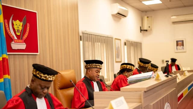 Cinq juges de la Cour constitutionnelle de la RDC, en toge rouge et toque bordée de peau de léopard, siègent en audience publique ; l'un d'eux lit une décision au micro, devant les armoiries et le drapeau congolais.