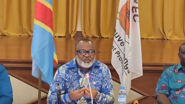 Corneille Nangaa, barbe blanche et lunettes, en chemise bleue imprimée, s'exprime devant un micro à une table de conférence de presse hérissée de micros de télévisions, entre le drapeau de la République démocratique du Congo et une bannière de l'Alliance Fleuve Congo.