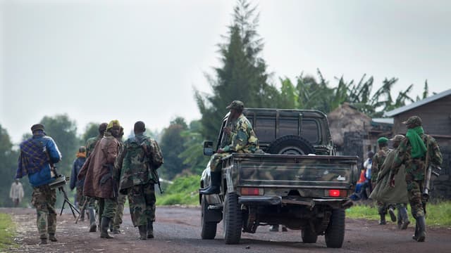 Une colonne de combattants armés en tenue de camouflage marche le long d'une route de terre, à côté d'un pick-up militaire, dans une zone rurale du Nord-Kivu ; des civils se tiennent en bordure de route.