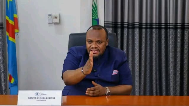 Daniel Bumba Lubaki, gouverneur de la ville de Kinshasa, assis à son bureau, la main levée, devant un chevalet portant son nom et son titre, entre le drapeau de la RDC et celui de la ville de Kinshasa.