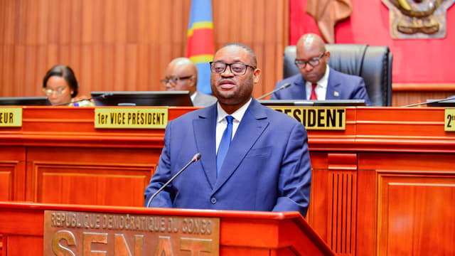 Le ministre congolais des Finances Doudou Fwamba, en costume bleu, s'exprime debout au pupitre marqué « République démocratique du Congo ,  Sénat », devant les membres du bureau assis à la tribune.