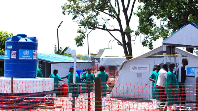 Une unité de traitement du choléra en République démocratique du Congo : des tentes blanches marquées « Organisation mondiale de la Santé » et « World Health Organization », des soignants en tenue verte et bottes blanches, une citerne d'eau bleue frappée du logo de l'OMS et « UK aid », le tout entouré d'une barrière de sécurité orange sur un terrain sablonneux.