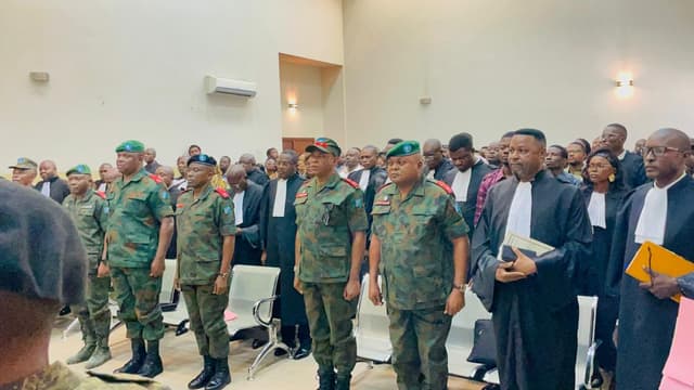 Des officiers généraux et supérieurs des FARDC en tenue de camouflage, debout dans la salle d'audience de la Haute Cour militaire, entourés d'avocats en robe noire et d'un public nombreux