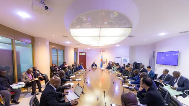 Vue d'ensemble d'une salle de réunion du ministère des Mines à Kinshasa, où des dizaines de représentants de sociétés minières sont assis autour d'une grande table ovale, devant le drapeau congolais et le portrait du chef de l'État.