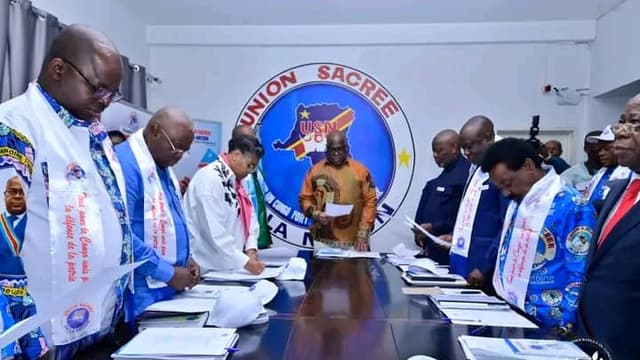 Des cadres de l'Union sacrée de la nation, écharpes aux couleurs de la plateforme, debout autour d'une table de réunion devant le logo de l'USN, à Kinshasa