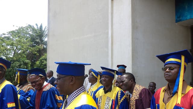 Un groupe de professeurs d'université en toge bleue, jaune et rouge, coiffés de la toque académique, réunis devant un bâtiment de l'Université de Kinshasa.