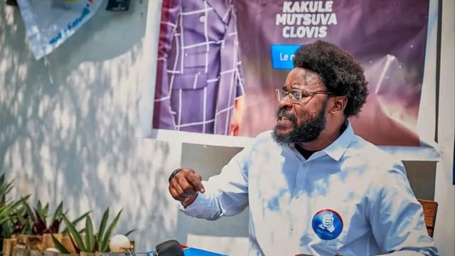 Clovis Mutsuva, en chemise bleu clair portant un badge du parti Ensemble pour la République, s'exprime devant plusieurs micros de presse, devant une banderole à son nom.