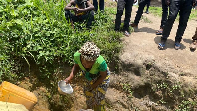 Une femme accroupie remplit des bidons jaunes avec une cuvette en puisant l'eau d'une source non aménagée creusée à même la terre, entourée de personnes debout au bord du trou.