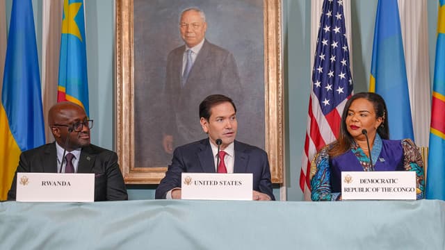 Trois responsables assis derrière une table recouverte d'une nappe bleue, devant des drapeaux du Rwanda, des États-Unis et de la République démocratique du Congo. Des plaques nominatives posées devant eux indiquent « Rwanda », « United States » et « Democratic Republic of the Congo ». À gauche, un homme en costume sombre portant des lunettes ; au centre, un homme en costume à cravate rouge qui parle dans un micro ; à droite, une femme en veste bleue et tenue en pagne coloré. Un portrait peint est accroché au mur derrière eux.