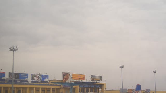 L'aérogare de l'aéroport international de N'Djili à Kinshasa, un long bâtiment jaune portant l'inscription « aéroport de ndjili » sur sa façade, avec plusieurs avions et des agents de piste stationnés sur le tarmac au premier plan.