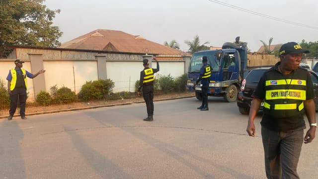 Des agents de la CNPR en gilets fluo régulent la circulation et un camion-benne sur une artère de Kamina, devant une villa au mur de clôture.
