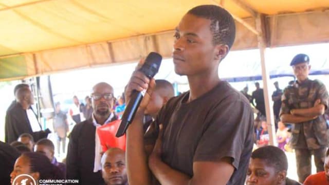 Un jeune homme debout, un micro à la main, répond aux questions du tribunal militaire de garnison de Lubumbashi lors d'une audience foraine sous une tente ; des avocats en robe noire et un militaire en béret bleu l'entourent.
