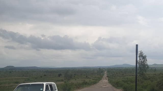 Route bitumée traversant la plaine de la Ruzizi, dans le territoire d'Uvira, avec une camionnette blanche à gauche, des collines à l'horizon et un ciel nuageux.