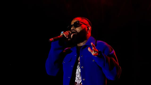 Le chanteur congolais Ferré Gola, veste bleue et lunettes de soleil, chante au micro sous des projecteurs rouges sur une scène de concert.