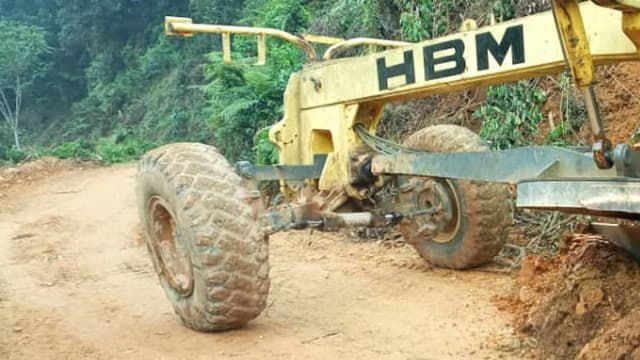 Une niveleuse jaune de marque HBM, aux gros pneus couverts de boue, travaille sur une route de terre fraîchement ouverte, taillée à flanc de colline à travers la forêt dense ; la terre ocre retournée s'entasse sur le bas-côté.