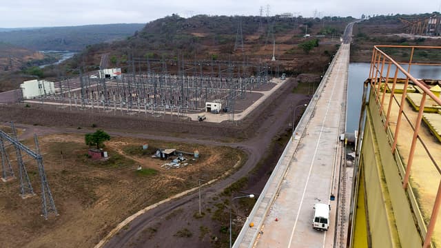 Vue aérienne d'un poste électrique à haute tension en Angola : rangées de portiques et d'appareillage métalliques, pylônes et lignes électriques s'éloignant vers des collines, le long de la crête d'un barrage hydroélectrique surplombant une retenue d'eau.