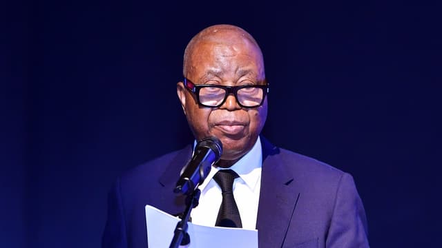 Adolphe Muzito, vice-Premier ministre au Budget, lit son discours au micro, une feuille à la main, sur une scène sombre marquée du logo de la Vice-Primature / Ministère du Budget.