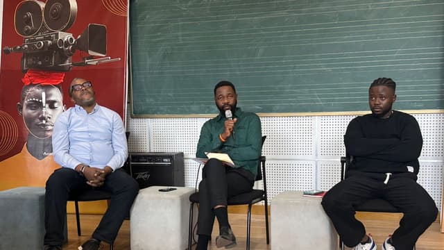 Trois organisateurs du FICKIN assis devant une affiche du festival lors de la conférence de presse de la 13e édition à Kinshasa, l'un d'eux parlant au micro
