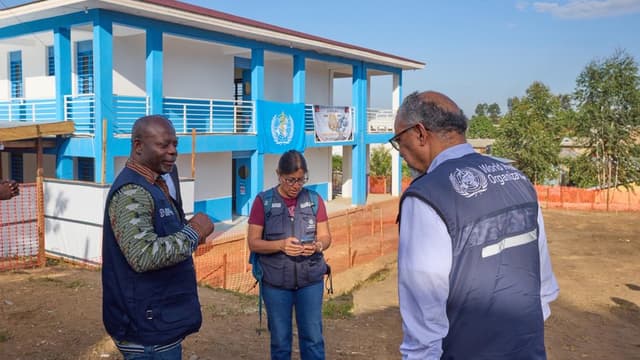 Deux responsables de la riposte et une agente de l'OMS en gilet bleu échangent dans la cour devant le bâtiment bleu et blanc du centre de traitement Ebola de Bunia, en Ituri, où flotte une banderole de l'OMS.