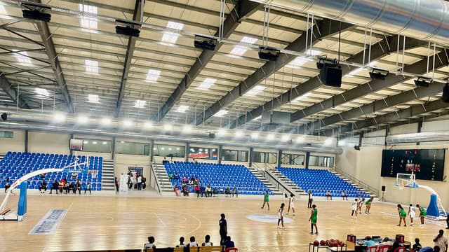 Vue intérieure du gymnase jumelé du Stade des Martyrs à Kinshasa : parquet de jeu clair, tribunes de sièges bleus partiellement occupées, tableau d'affichage électronique et charpente métallique de la halle, pendant une rencontre.