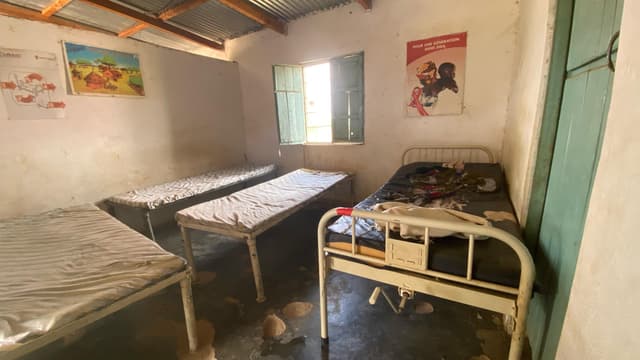 Salle de soins désertée d'une structure de santé aux murs blanchis : trois lits métalliques et une table d'examen vides, sol maculé, affiches sanitaires (USAID, « Pour une génération sans sida ») décollées aux murs, fenêtre à volets de bois entrouverte.