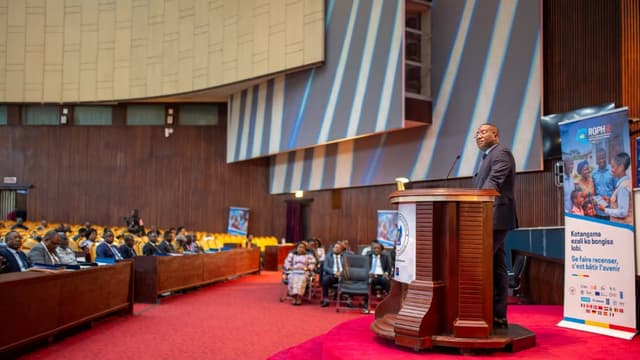 Guylain Nyembo s'exprime depuis un pupitre dans l'hémicycle du Palais du Peuple à Kinshasa, devant une assistance assise, à côté d'un kakémono du RGPH-2 portant le slogan « Se faire recenser, c'est bâtir l'avenir »