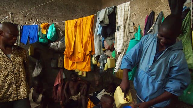 Intérieur d'une cellule surpeuplée de la prison de Bunia : des détenus assis et allongés à même le sol en terre battue, du linge suspendu à une corde le long du mur en béton, des bidons jaunes et des bouteilles d'eau accrochés à la paroi. Un homme debout verse de l'eau d'un bidon dans une tasse bleue.