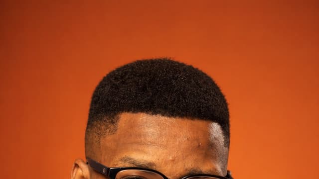Portrait d'Angello Luvungu Muhiya, en veste bordeaux et col roulé noir, sur fond orange.
