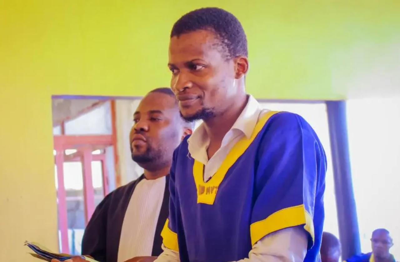 Nathanaël Onokomba, vêtu de la tenue bleu et jaune des détenus, se tient debout aux côtés d'un avocat en robe noire dans une salle aux murs verts.