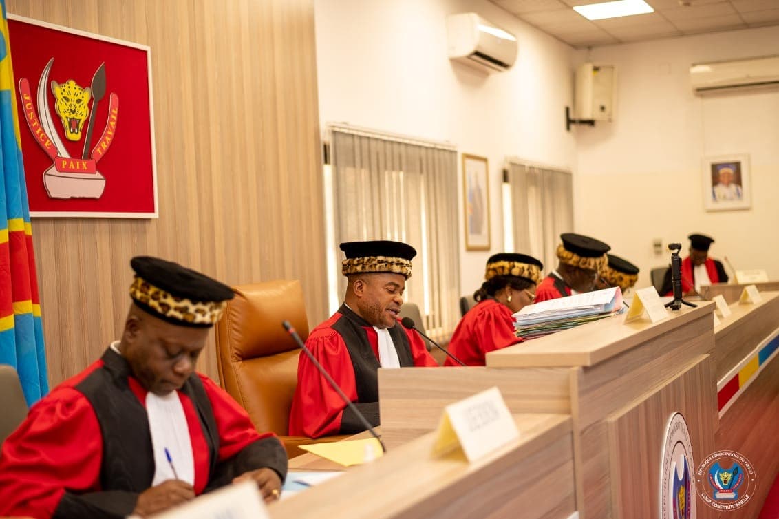 Cinq juges de la Cour constitutionnelle de la RDC, en toge rouge et toque bordée de peau de léopard, siègent en audience publique ; l'un d'eux lit une décision au micro, devant les armoiries et le drapeau congolais.