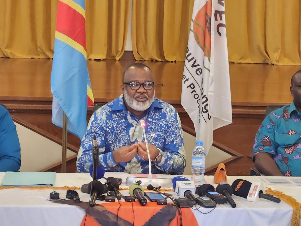 Corneille Nangaa, barbe blanche et lunettes, en chemise bleue imprimée, s'exprime devant un micro à une table de conférence de presse hérissée de micros de télévisions, entre le drapeau de la République démocratique du Congo et une bannière de l'Alliance Fleuve Congo.