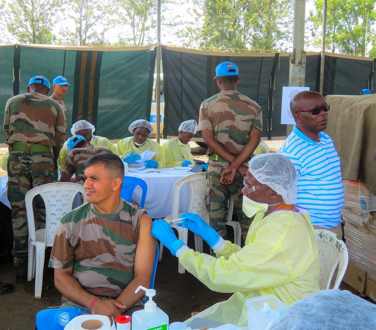 Séance de vaccination contre Ebola en plein air en République démocratique du Congo : des agents de santé congolais en blouse jaune, gants bleus, masque et charlotte administrent un vaccin dans le bras d'un homme assis, tandis que d'autres soignants enregistrent les personnes vaccinées à une table.