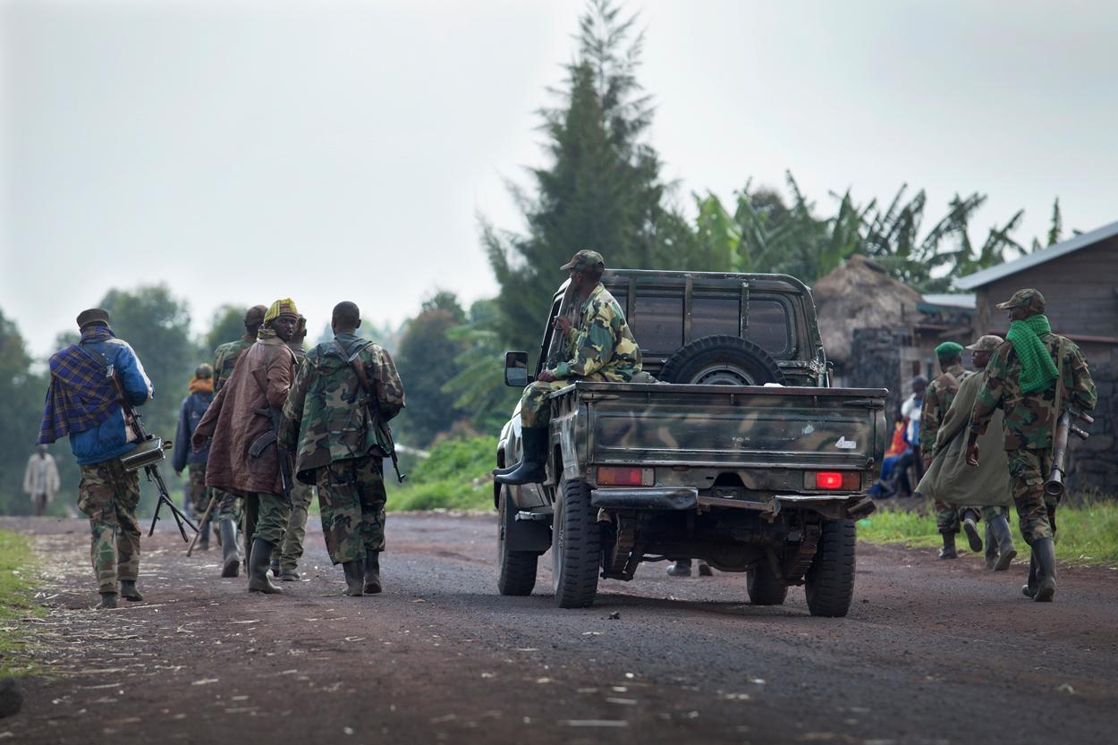 Une colonne de combattants armés en tenue de camouflage marche le long d'une route de terre, à côté d'un pick-up militaire, dans une zone rurale du Nord-Kivu ; des civils se tiennent en bordure de route.