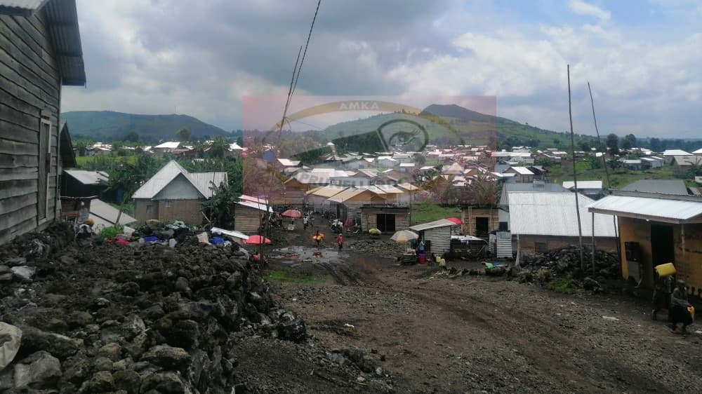Vue d'une localité du territoire de Nyiragongo : maisons aux toits de tôle bâties sur un sol de roche volcanique, le long d'une route de terre, avec les collines des Virunga en arrière-plan.