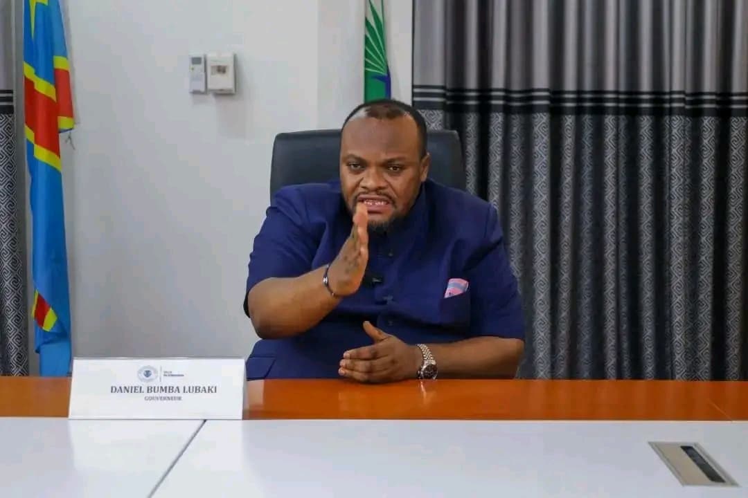 Daniel Bumba Lubaki, gouverneur de la ville de Kinshasa, assis à son bureau, la main levée, devant un chevalet portant son nom et son titre, entre le drapeau de la RDC et celui de la ville de Kinshasa.