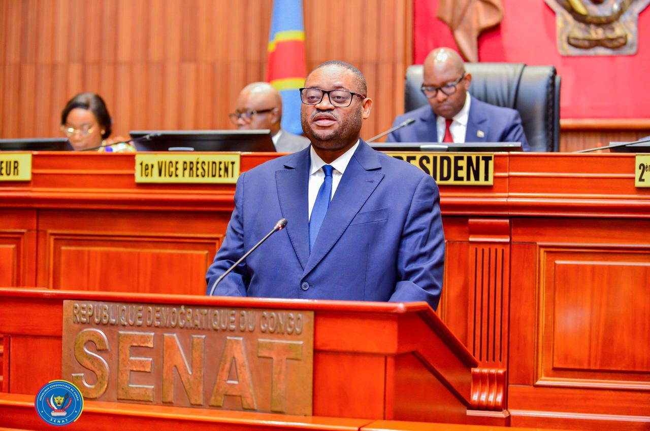 Le ministre congolais des Finances Doudou Fwamba, en costume bleu, s'exprime debout au pupitre marqué « République démocratique du Congo ,  Sénat », devant les membres du bureau assis à la tribune.