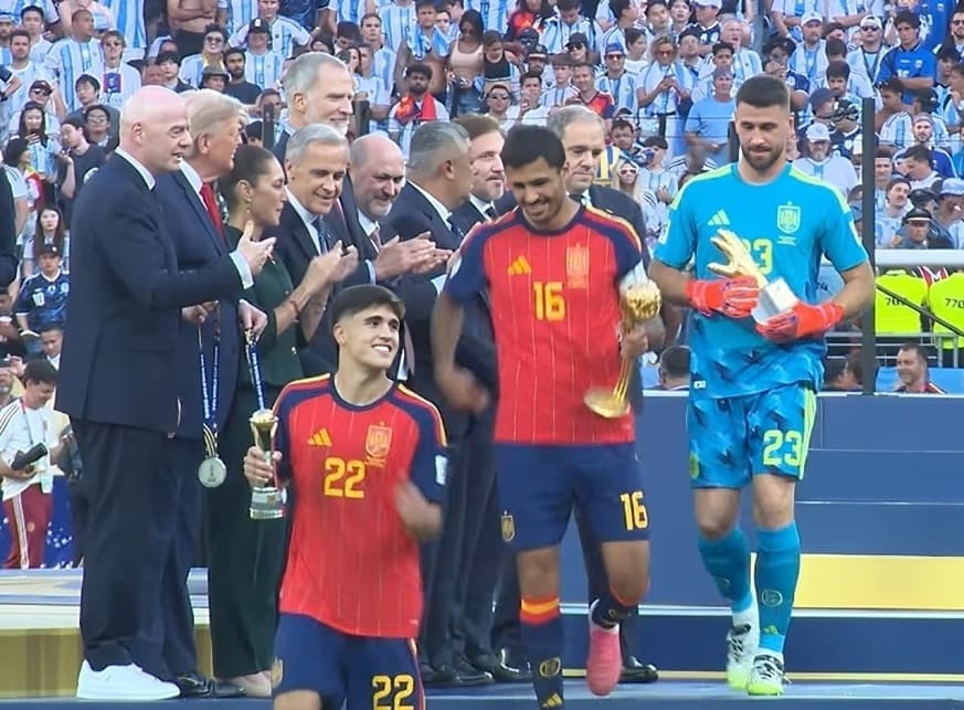 Sur le podium du MetLife Stadium, Rodri, maillot rouge numéro 16, tient le trophée du Ballon d'or à côté de Pau Cubarsi, numéro 22, et du gardien Unai Simon en tenue bleue, devant Gianni Infantino et les officiels qui applaudissent.