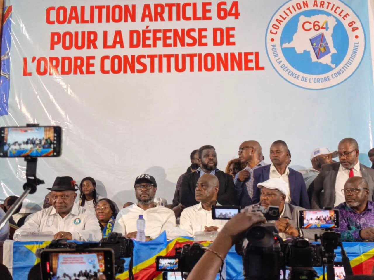 Les responsables de la Coalition Article 64 assis derrière une table drapée du drapeau congolais, devant une grande banderole blanche portant en lettres rouges « Coalition Article 64 pour la défense de l'ordre constitutionnel » et le logo C64 ; au premier plan, des caméras et des téléphones filment la séance.