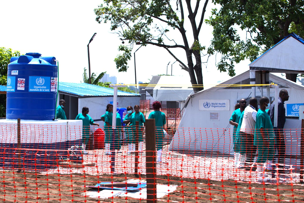 Une unité de traitement du choléra en République démocratique du Congo : des tentes blanches marquées « Organisation mondiale de la Santé » et « World Health Organization », des soignants en tenue verte et bottes blanches, une citerne d'eau bleue frappée du logo de l'OMS et « UK aid », le tout entouré d'une barrière de sécurité orange sur un terrain sablonneux.
