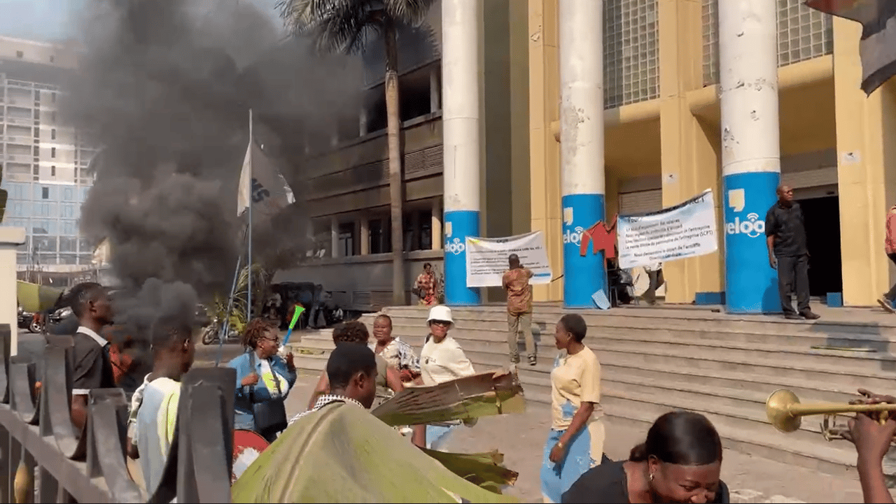 Agents de la SCPT en grève devant le siège de l'entreprise à Kinshasa : fumée noire de pneus brûlés, banderoles réclamant le départ de la direction, manifestants avec vuvuzelas et trompette.