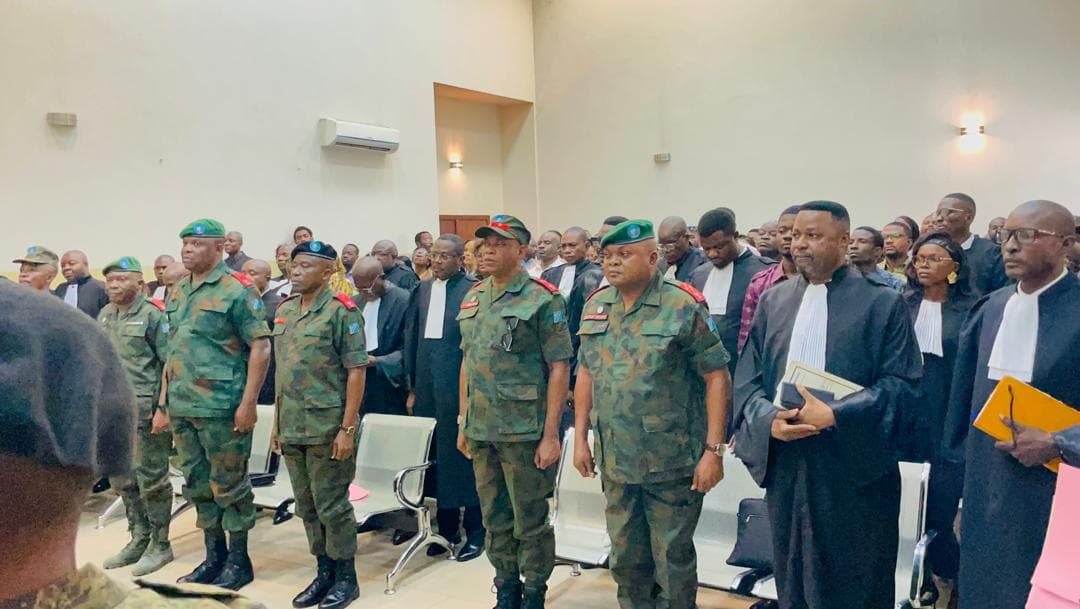 Des officiers généraux et supérieurs des FARDC en tenue de camouflage, debout dans la salle d'audience de la Haute Cour militaire, entourés d'avocats en robe noire et d'un public nombreux